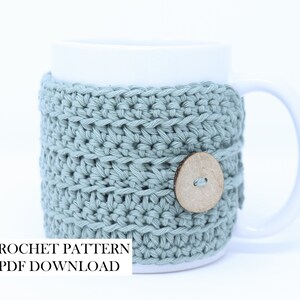 May include: A white ceramic mug featuring a sage green crocheted cosy with a round wooden button closure. The text CROCHET PATTERN PDF DOWNLOAD is printed in black on the lower left side of the image, indicating a digital file for this item.