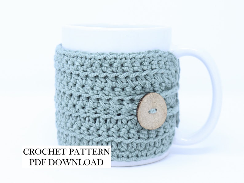 May include: A white ceramic mug featuring a sage green crocheted cosy with a round wooden button closure. The text CROCHET PATTERN PDF DOWNLOAD is printed in black on the lower left side of the image, indicating a digital file for this item.