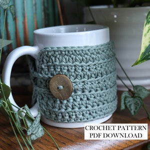 May include: A white ceramic mug wrapped in a sage green crocheted cosy featuring a single round wooden button closure. The text CROCHET PATTERN PDF DOWNLOAD is printed in black on a white rectangular background at the bottom of the image.