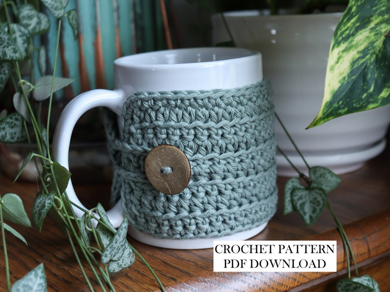 May include: A white ceramic mug wrapped in a sage green crocheted cosy featuring a single round wooden button closure. The text CROCHET PATTERN PDF DOWNLOAD is printed in black on a white rectangular background at the bottom of the image.