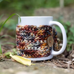 May include: A white ceramic mug featuring a removable, hand-crocheted cosy in a variegated yarn pattern of deep red, brown, grey, and gold tones. The cosy is secured around the mug with a single, round, light brown wooden button on the side.