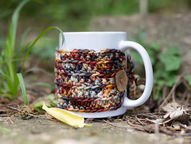 May include: A white ceramic mug featuring a removable, hand-crocheted cosy in a variegated yarn pattern of deep red, brown, grey, and gold tones. The cosy is secured around the mug with a single, round, light brown wooden button on the side.