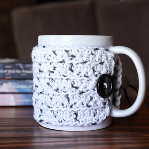 May include: A white ceramic mug featuring a removable, hand-crocheted cosy in a white yarn with black flecks. The cosy wraps around the mug and is secured on the side with a single, round, black button, providing a textured layer over the smooth surface.