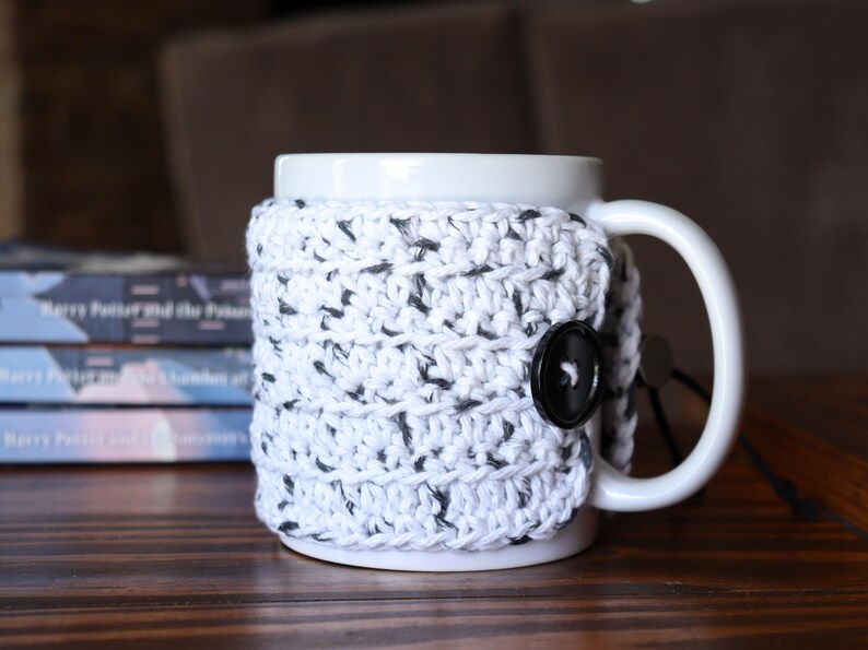 May include: A white ceramic mug featuring a removable, hand-crocheted cosy in a white yarn with black flecks. The cosy wraps around the mug and is secured on the side with a single, round, black button, providing a textured layer over the smooth surface.