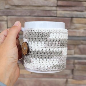 May include: A white ceramic mug featuring a removable crochet cosy. The cosy is crafted with alternating horizontal stripes of light grey and white yarn, secured on the side with a single round, dark brown wooden button with a visible centre hole.