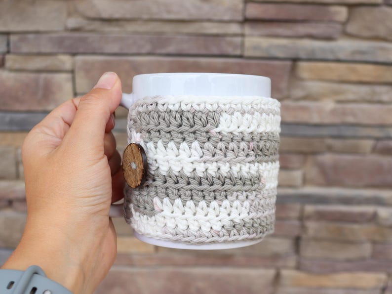 May include: A white ceramic mug featuring a removable crochet cosy. The cosy is crafted with alternating horizontal stripes of light grey and white yarn, secured on the side with a single round, dark brown wooden button with a visible centre hole.