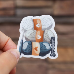 Pu&ograve; includere: Un adesivo con un'illustrazione di cartone animato di tre gomitoli di lana, due bianchi e uno blu, con un uncinetto. I gomitoli di lana sono impilati uno sopra l'altro. L'adesivo ha il testo "yarn addict" sul lato.