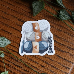 Pu&ograve; includere: Un adesivo con un'illustrazione di tre gomitoli di lana, due bianchi e uno blu, con un ferretto e la scritta "yarn addict".