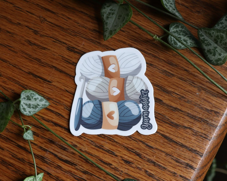 Pu&ograve; includere: Un adesivo con un'illustrazione di tre gomitoli di lana, due bianchi e uno blu, con un ferretto e la scritta "yarn addict".