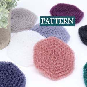 May include: A collection of seven crocheted scrubbies in various colors, including pink, purple, gray, white, black, and green. The scrubbies are round and have a textured surface. The word "PATTERN" is written in green text on a white background.