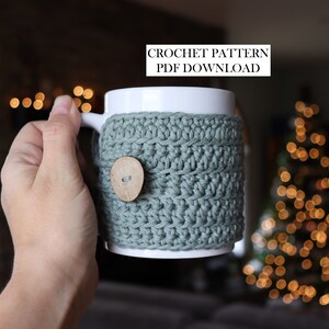 May include: A white ceramic mug featuring a sage green crocheted cosy with a single round wooden button. The text CROCHET PATTERN PDF DOWNLOAD is visible at the top. The cosy is textured with horizontal rows of yarn stitches wrapped around the mug.