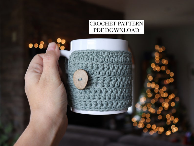 May include: A white ceramic mug featuring a sage green crocheted cosy with a single round wooden button. The text CROCHET PATTERN PDF DOWNLOAD is visible at the top. The cosy is textured with horizontal rows of yarn stitches wrapped around the mug.
