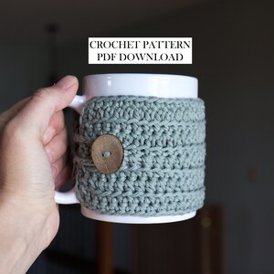 May include: A white ceramic mug featuring a sage green crocheted cosy with a round wooden button closure. The text CROCHET PATTERN PDF DOWNLOAD is visible above the mug. The cosy is textured with horizontal rows of yarn stitches.