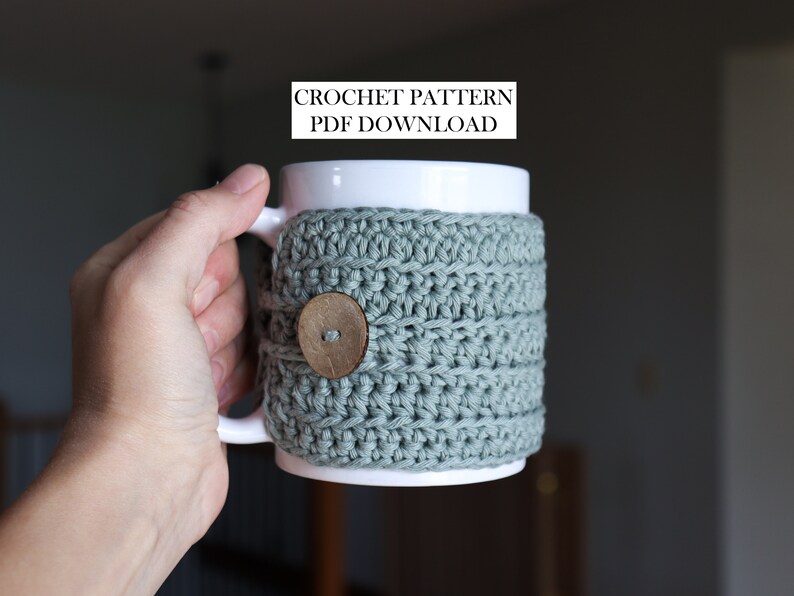 May include: A white ceramic mug featuring a sage green crocheted cosy with a round wooden button closure. The text CROCHET PATTERN PDF DOWNLOAD is visible above the mug. The cosy is textured with horizontal rows of yarn stitches.