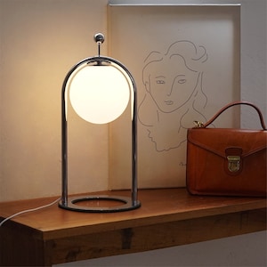 May include: A chrome table lamp with a white globe shade. The lamp has a curved arch and a round base. The lamp is on a wooden table next to a brown leather handbag and a framed drawing.