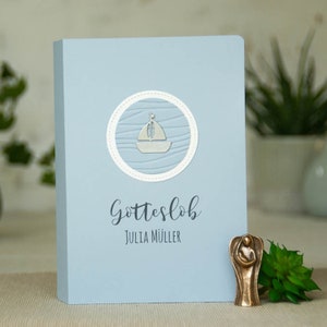 Könnte beinhalten: Eine hellblaue Holzkiste mit einem weißen Kreis, der ein silbernes Segelboot auf einem blau-weißen Wellenmuster enthält. Der Text "Gotteslob" und "Julia Müller" ist in Schwarz unter dem Kreis gedruckt.