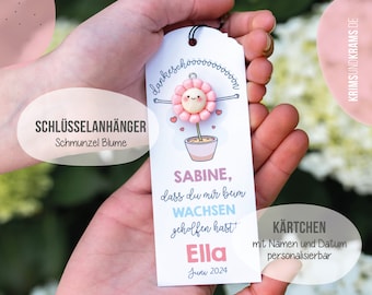 May include: A pink and white flower-shaped keychain with a smiling face. The keychain is attached to a card that says "Sabine, dass du mir beim Wachsen geholfen hast Ella Juni 2024". The card also says "Schlüsselanhänger Schmunzel Blume" and "KÄRTCHEN mit Namen und Datum personalisierbar".