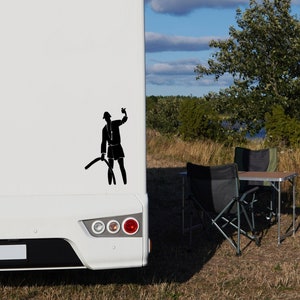 May include: A white recreational vehicle featuring a black vinyl decal on the rear panel. The decal depicts a silhouette of a person wearing a hat, holding a small object in one hand and a long, curved object in the other, positioned near the taillights.