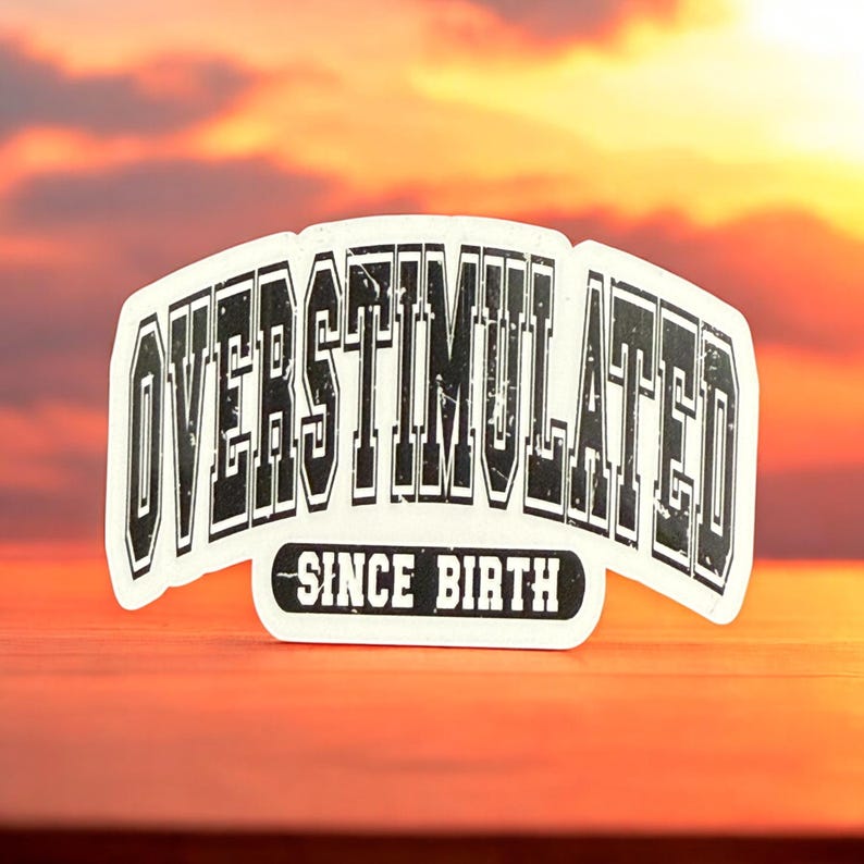 May include: A white sticker featuring the text "OVERSTIMULATED" in a bold, black, distressed font. Beneath, the words "SINCE BIRTH" are in a smaller, black font. The sticker is set against a warm, sunset-coloured background.