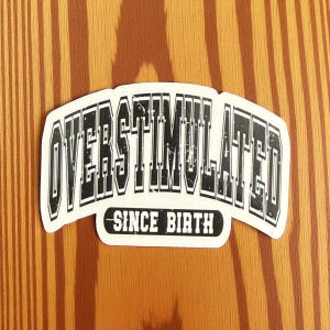 May include: A white sticker with the words "OVERSTIMULATED" in bold, black, block letters, arched across the top. Below, in a smaller, black, rectangular box, the words "SINCE BIRTH" are printed. The sticker is set against a wood grain background.