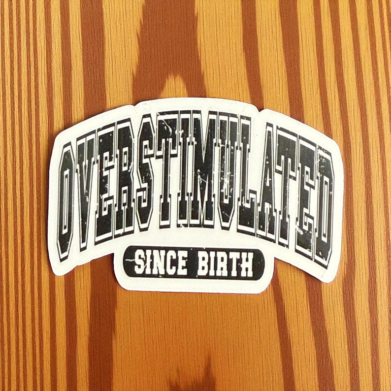 May include: A white sticker with the words "OVERSTIMULATED" in bold, black, block letters, arched across the top. Below, in a smaller, black, rectangular box, the words "SINCE BIRTH" are printed. The sticker is set against a wood grain background.