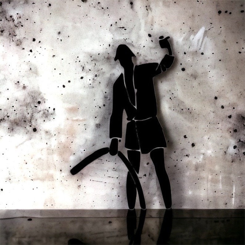 May include: A black silhouette wall art piece depicting a figure wearing a hat and coat, holding a curved object in one hand and raising the other hand. The figure is set against a light gray textured background with dark speckles and spots.