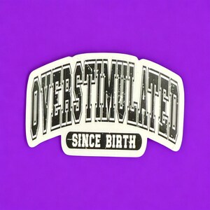 May include: A white sticker with the words "OVERSTIMULATED" in a bold, distressed font, arched across the top. Below, the text "SINCE BIRTH" is centered. The sticker is set against a vibrant purple background.