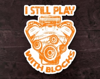 May include: A die-cut sticker featuring a distressed orange graphic of a V8 engine block. The text I STILL PLAY WITH BLOCKS is printed in a bold, distressed orange font above and below the engine illustration on a white background.