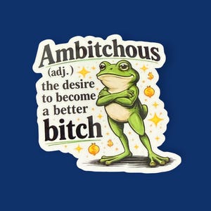 Op de afbeelding: Een sticker met een illustratie van een groene kikker en de tekst "Ambitchous (adj.) de wens om een betere bitch te worden." De kikker heeft zijn armen gekruist en is omringd door gouden sterren en dollartekens.