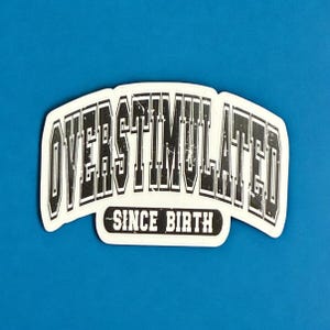 May include: A white sticker with the words "OVERSTIMULATED" in a bold, black, distressed font. Below, in a smaller font, it reads "SINCE BIRTH". The sticker has a curved shape and is set against a blue background.