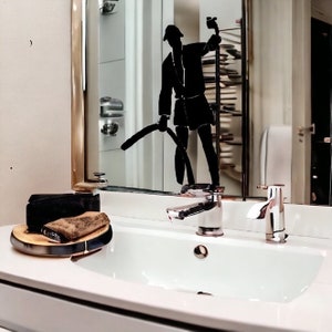 May include: A white bathroom sink with a chrome faucet and a gold-trimmed mirror. A black silhouette decal of a person holding a vacuum cleaner is applied to the mirror surface. A wooden tray with a black object sits on the white countertop.
