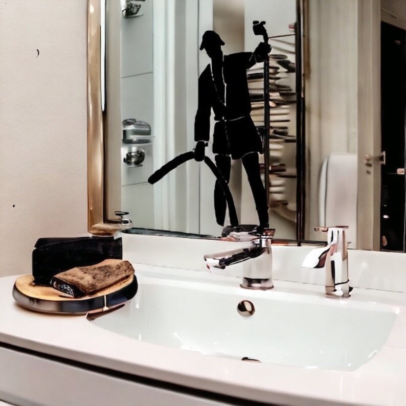 May include: A white bathroom sink with a chrome faucet and a gold-trimmed mirror. A black silhouette decal of a person holding a vacuum cleaner is applied to the mirror surface. A wooden tray with a black object sits on the white countertop.