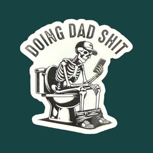 Op de afbeelding: Een zwart-witte sticker met een skelet met een pet en een smartphone op een toilet. De sticker heeft de tekst "DOING DAD SHIT" in een witte, afgeronde letter.