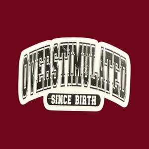 May include: A white sticker with the words "OVERSTIMULATED" in a bold, black, distressed font, arched across the top. Below, in a smaller font, it reads "SINCE BIRTH". The sticker is set against a dark red background.