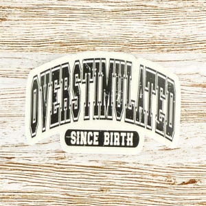 May include: A white sticker featuring the text "OVERSTIMULATED" in a bold, black, arched font. Beneath, in a smaller, black font, it reads "SINCE BIRTH". The sticker is set against a light wood grain background.