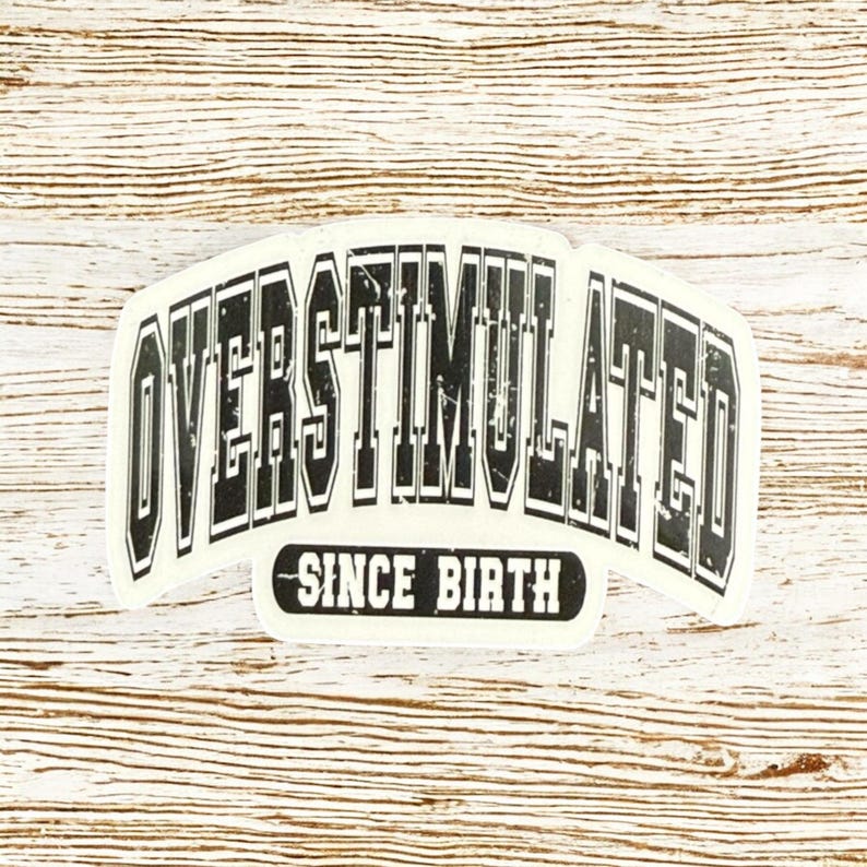 May include: A white sticker featuring the text "OVERSTIMULATED" in a bold, black, arched font. Beneath, in a smaller, black font, it reads "SINCE BIRTH". The sticker is set against a light wood grain background.