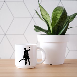 May include: A white ceramic mug featuring a black silhouette graphic of a person wearing a hat and holding a curved object. The mug has a smooth, cylindrical body with a side handle and a clean, minimalist white finish throughout the exterior.