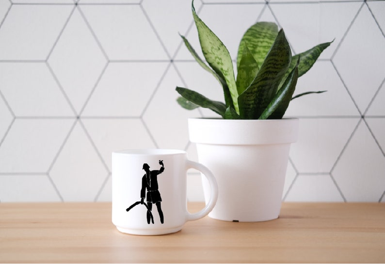 May include: A white ceramic mug featuring a black silhouette graphic of a person wearing a hat and holding a curved object. The mug has a smooth, cylindrical body with a side handle and a clean, minimalist white finish throughout the exterior.