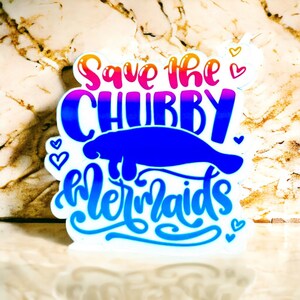 Peut inclure: Un autocollant blanc avec un lamantin bleu et le texte "Save the Chubby Mermaids" en couleurs arc-en-ciel. L'autocollant a de petits c&oelig;urs autour du texte.