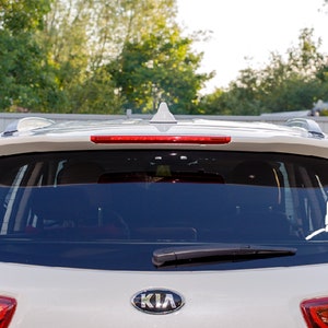 May include: A white vinyl decal applied to the rear window of a light-colored vehicle. The decal features a silhouette of a firefighter in uniform holding an axe in one hand and a fire hose in the other, positioned on the right side of the glass.