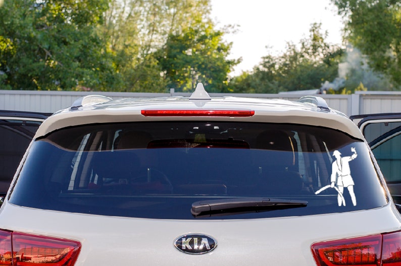 May include: A white vinyl decal applied to the rear window of a light-colored vehicle. The decal features a silhouette of a firefighter in uniform holding an axe in one hand and a fire hose in the other, positioned on the right side of the glass.