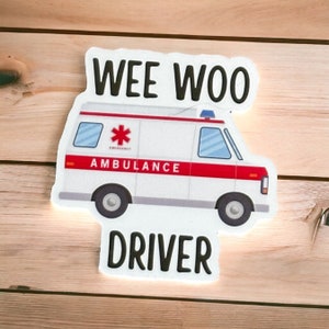 Op de afbeelding: Witte ambulance sticker met een rode streep en de woorden "WEE WOO DRIVER" in het zwart. De ambulance heeft een rood kruis en het woord "AMBULANCE" aan de zijkant.