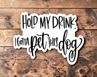 Könnte beinhalten: Ein weißer, konturgestanzter Aufkleber mit schwarzer, handschriftlicher Typografie, die den Text "HOLD MY DRINK. I GOTTA PET THIS DOG." zeigt. Der Text ist in zwei Zeilen angeordnet und kombiniert Blockschrift mit kursiver Schrift auf weißem Grund.