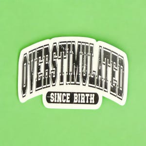 May include: A white sticker with the words "OVERSTIMULATED" in a bold, black, distressed font, arched across the top. Below, in a smaller, black font, it reads "SINCE BIRTH". The sticker is set against a bright green background.