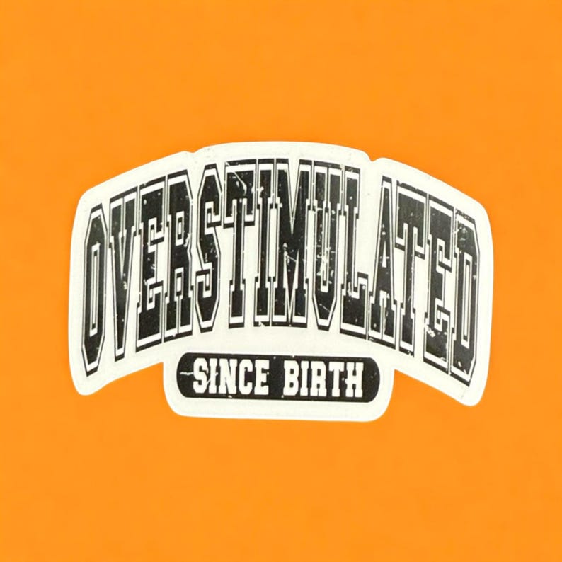 May include: A white sticker with the words "OVERSTIMULATED" in a bold, distressed font, arched across the top. Below, the text "SINCE BIRTH" is in a smaller, curved font. The sticker is set against a bright orange background.