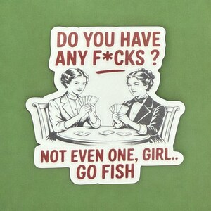 May include: A white sticker with a red border and text. The sticker features two women playing cards with the text "DO YOU HAVE ANY F*CKS? NOT EVEN ONE, GIRL.. GO FISH."