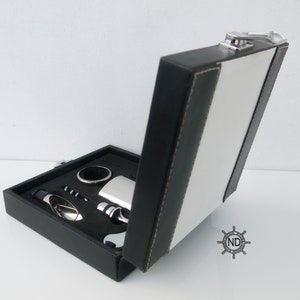 Peut inclure: Une boîte cadeau en similicuir noir avec un fermoir argenté, ouverte pour révéler un ensemble d'accessoires à vin. L'ensemble comprend un tire-bouchon, un bouchon de bouteille, un verseur et une petite fiole.