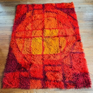 May include: A rectangular shag rug with a vibrant, retro design. The rug features a bold color palette of red, orange, and yellow, with a geometric pattern in the center. The rug is placed on a wooden floor.