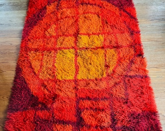 May include: A rectangular shag rug with a vibrant, retro design. The rug features a bold color palette of red, orange, and yellow, with a geometric pattern in the center. The rug is placed on a wooden floor.