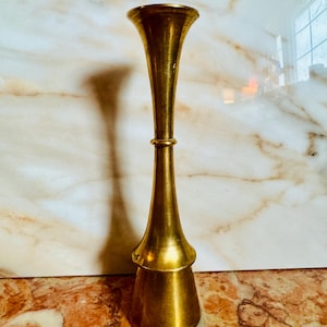 May include: A tall, brass-colored vase with a flared top and base, connected by a slender, cylindrical midsection. The vase has a tiered design and stands approximately 12 inches tall. The smooth, polished surface reflects light, creating highlights and shadows.